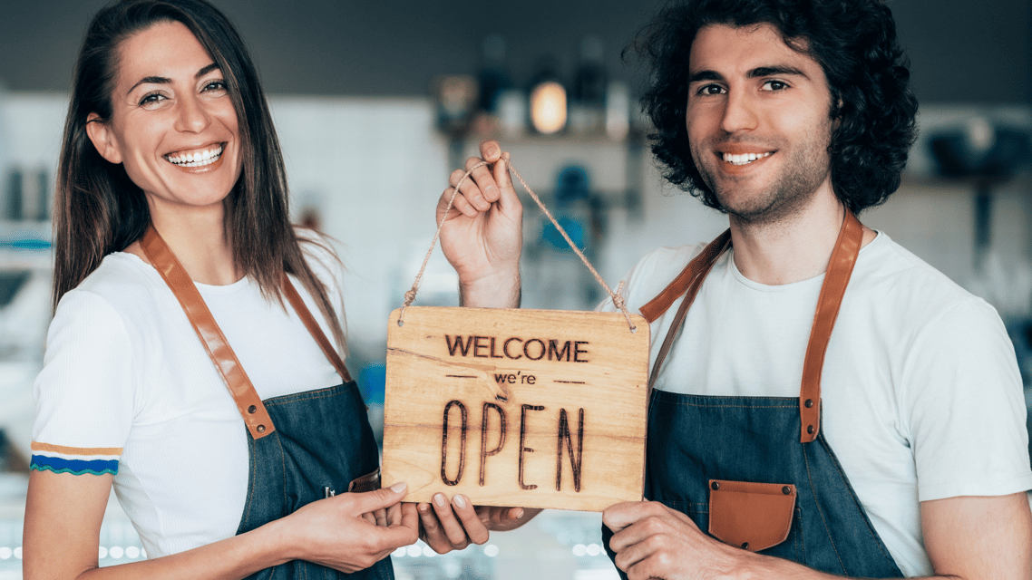 man and woman holding wooden sign with text saying welcome we are open